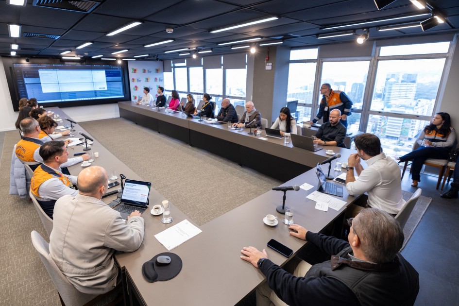 A fotografia em plano geral e ângulo ligeiramente picado (de cima para baixo) registra uma reunião de trabalho institucional de alto nível em uma sala de conferências moderna, identificada pelo título do arquivo como parte da estratégia de preparação para eventos climáticos.
Disposição e Participantes
A Grande Mesa: Os participantes (cerca de 20 pessoas) estão distribuídos em torno de uma grande mesa de reuniões em formato de "U" na cor cinza-escura.
Equipes Técnicas: No lado esquerdo, em primeiro plano, destacam-se integrantes vestindo coletes oficiais bicolores (laranja na parte superior e azul-marinho na inferior) com a inscrição "DEFESA CIVIL" nas costas, indicando a liderança ou forte presença do órgão no encontro.
Ferramentas de Trabalho: Sobre a mesa, veem-se diversos notebooks, tablets abertos com teclados acoplados, microfones de mesa com pedestais baixos, folhas de papel com pautas, além de xícaras de café e copos de água individuais.
O Ambiente e Recursos Visuais
Apresentação Central: Fixada na parede ao fundo, à esquerda, há uma grande tela de TV ou painel digital que exibe uma interface de sistema operacional com tabelas e dados técnicos em tons de azul.
Identidade Visual: Próximo à tela ao fundo, um painel vertical branco exibe logotipos coloridos repetidos, que remetem à identidade visual do governo do estado.
Infraestrutura e Iluminação: O teto da sala é preto com trilhos de spots de luz LED direcionais acesos. O chão é revestido com carpete cinza-claro na área central da mesa. À direita, grandes janelas de vidro que vão do chão ao teto revelam uma vista panorâmica para o horizonte urbano de uma cidade em um dia nublado.