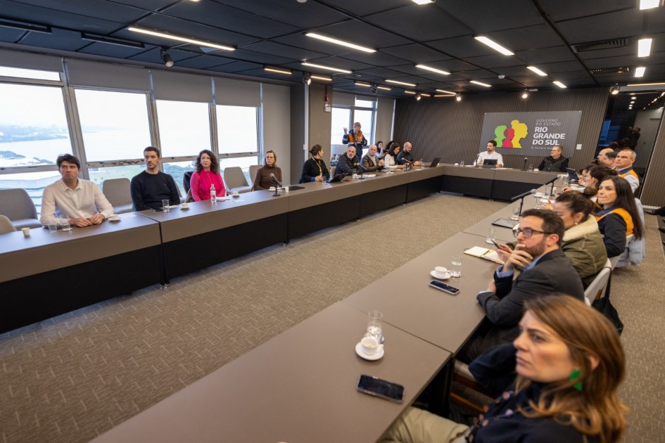 A fotografia em plano geral e ângulo ligeiramente picado registra uma reunião de trabalho institucional com diversos gestores e técnicos, identificada pelo título do arquivo como parte de uma estratégia estadual de redução de riscos a eventos extremos.
Disposição e Participantes
A Grande Mesa: Os participantes (cerca de 20 pessoas) estão dispostos ao redor de uma grande mesa cinza-escura em formato de