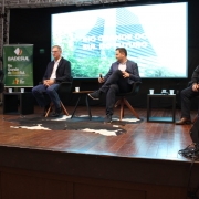 Esta &eacute; uma descri&ccedil;&atilde;o da imagem para fins de acessibilidade:

A fotografia em plano m&eacute;dio e &acirc;ngulo frontal registra um painel de debate ocorrendo no palco de um audit&oacute;rio, identificado pelo nome do arquivo como a participa&ccedil;&atilde;o do Governo do Estado no F&oacute;rum de Competitividade no Vale do Taquari.

Os Painelistas e a Disposi&ccedil;&atilde;o
Participantes: Quatro homens est&atilde;o sentados em poltronas pretas individuais com p&eacute;s de madeira, dispostas em linha ao longo do palco.

Composi&ccedil;&atilde;o (da esquerda para a direita):

O primeiro homem, vestindo terno escuro e camisa branca, est&aacute; ligeiramente inclinado para a frente e fala ao microfone.

O segundo homem usa um blazer escuro, camisa clara e cal&ccedil;a jeans, mantendo uma postura de escuta com as m&atilde;os cruzadas no colo.

O terceiro homem, com as pernas cruzadas, veste blazer escuro e cal&ccedil;a escura, segurando um microfone enquanto gesticula com a outra m&atilde;o.

O quarto homem, posicionado na extrema direita, usa &oacute;culos, uma blusa de gola alta clara sob uma jaqueta escura e cal&ccedil;a escura.

Elementos de Cenografia e Proje&ccedil;&atilde;o
Tel&atilde;o de Fundo: Uma grande tela digital centralizada ao fundo exibe uma imagem de um edif&iacute;cio moderno de arquitetura marcante e vegeta&ccedil;&atilde;o ao redor. Sobreposta &agrave; imagem, h&aacute; a inscri&ccedil;&atilde;o em letras mai&uacute;sculas brancas: "RIO GRANDE DO SUL DO FUTURO".

Banner Institucional: &Agrave; esquerda, h&aacute; um banner vertical verde com o logotipo e o nome do "BADESUL DESENVOLVIMENTO", seguido da frase "Rio Grande do Badesul".

Decora&ccedil;&atilde;o do Palco: O ch&atilde;o do palco &eacute; feito de t&aacute;buas de madeira compridas dispostas horizontalmente. No centro, sob as duas poltronas centrais, h&aacute; um grande tapete de couro bovino com manchas pretas e brancas. Pequenas mesas de apoio pretas com copos de &aacute;gua est&atilde;o posicionadas entre os participantes.

Ambiente e Detalhes T&eacute;cnicos
Estrutura: O palco &eacute; emoldurado por uma estrutura met&aacute;lica de treli&ccedil;a (box truss) vis&iacute;vel no canto superior esquerdo e por cortinas pretas ao fundo.

Equipamentos: No canto inferior direito, em primeiro plano, destaca-se uma caixa de retorno de som preta voltada para o palco, com alguns fios conectados.