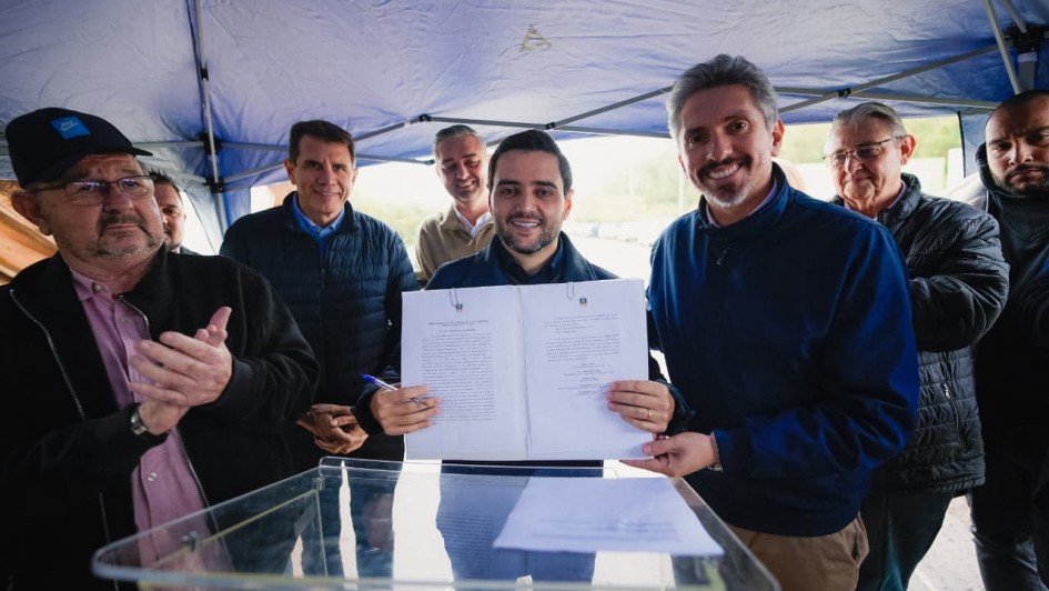 Grupo de pessoas reunidas sob uma tenda ao ar livre, onde duas pessoas ao centro seguram um documento aberto voltado para a c&acirc;mera, enquanto outras ficam ao redor pr&oacute;ximas a um p&uacute;lpito transparente.