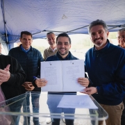 Grupo de pessoas reunidas sob uma tenda ao ar livre, onde duas pessoas ao centro seguram um documento aberto voltado para a c&acirc;mera, enquanto outras ficam ao redor pr&oacute;ximas a um p&uacute;lpito transparente.