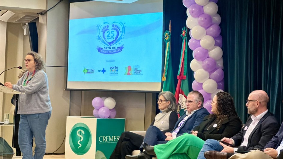 A fotografia em plano m&eacute;dio e &acirc;ngulo frontal registra a realiza&ccedil;&atilde;o de uma cerim&ocirc;nia institucional em um audit&oacute;rio, identificado pelo nome do arquivo e pelas proje&ccedil;&otilde;es visuais como o "Evento de 25 anos da triagem neonatal 2026_1.jpeg".

A Palestrante e a Mesa de Autoridades
A&ccedil;&atilde;o Principal: No lado esquerdo, em p&eacute; atr&aacute;s de um p&uacute;lpito transparente, uma mulher de &oacute;culos, cabelos cacheados e vestindo um casaco de l&atilde; cinza com cal&ccedil;a jeans azul fala ao microfone direcionado. Ela usa um colar de contas coloridas.

Composi&ccedil;&atilde;o da Mesa: &Agrave; direita, dispostos em cadeiras estofadas, quatro integrantes (duas mulheres e dois homens) acompanham o discurso.

A primeira mulher veste uma blusa de gola alta clara e cal&ccedil;a escura.

O homem ao lado usa blazer escuro, camisa social clara e cal&ccedil;a verde vibrante.

A terceira integrante possui cabelos escuros cacheados e veste um casaco preto com cal&ccedil;a jeans azul.

O quarto homem, de &oacute;culos e cal&ccedil;a bege, est&aacute; com as pernas cruzadas e observa o evento. Na extremidade direita, nota-se o bra&ccedil;o de um quinto participante.

Elementos Visuais e Cenografia
Tel&atilde;o de Proje&ccedil;&atilde;o: Uma grande tela centralizada ao fundo exibe um slide azul claro com um logotipo comemorativo em formato de cora&ccedil;&atilde;o estilizado com pezinhos de beb&ecirc;, onde se l&ecirc; "25 ANOS / SRTN RS / TRIAGEM NEONATAL". Na parte inferior do slide, aparecem os logotipos dos realizadores, incluindo o da Prefeitura de Porto Alegre e o do Governo do Estado do Rio Grande do Sul.

Decora&ccedil;&atilde;o e Identidade: Abaixo da tela, h&aacute; um balc&atilde;o branco e verde com o logotipo do CREMERS (Conselho Regional de Medicina do Estado do Rio Grande do Sul), que traz o s&iacute;mbolo da medicina (a fita e a serpente) em um c&iacute;rculo verde. Pequenos arranjos e colunas de bal&otilde;es infl&aacute;veis nas cores branca e lil&aacute;s decoram o cen&aacute;rio.

O Ambiente: O palco possui piso claro e, &agrave; direita, h&aacute; uma cortina de fundo escura plissada. No canto inferior esquerdo, sobre o ch&atilde;o, veem-se garrafas de &aacute;gua dispostas para os participantes.