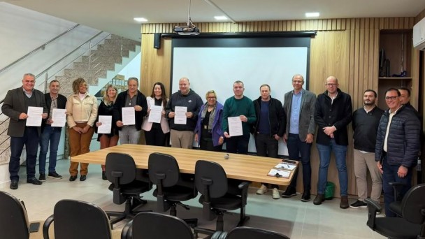 Esta &eacute; uma descri&ccedil;&atilde;o da imagem para fins de acessibilidade:

A fotografia em plano geral e &acirc;ngulo frontal registra um grupo de autoridades e gestores reunidos em uma sala de reuni&otilde;es, posando para uma foto institucional. O t&iacute;tulo do arquivo identifica o ato como a apresenta&ccedil;&atilde;o de a&ccedil;&otilde;es de prote&ccedil;&atilde;o &agrave;s mulheres pelo Governo do Estado.

O Grupo e a A&ccedil;&atilde;o
Participantes: Um grupo de 15 pessoas (10 homens e 5 mulheres) est&aacute; posicionado em p&eacute;, lado a lado, formando uma linha horizontal ao fundo da sala. Todos est&atilde;o voltados para a c&acirc;mera com express&otilde;es formais e sorridentes.

Exibi&ccedil;&atilde;o de Documentos: Sete pessoas localizadas na metade esquerda do grupo seguram folhas de papel branco com textos impressos (documentos oficiais ou termos de ades&atilde;o), exibindo-os de forma vis&iacute;vel para registrar a assinatura ou celebra&ccedil;&atilde;o de um ato.

O Ambiente e Mobili&aacute;rio
Mesa de Reuni&otilde;es: Em primeiro plano, h&aacute; uma longa mesa de reuni&otilde;es retangular de madeira clara. Ao redor dela, est&atilde;o dispostas cadeiras de escrit&oacute;rio pretas com rod&iacute;zios. Sobre a mesa, no canto direito, veem-se algumas pastas de vota&ccedil;&atilde;o ou pranchetas e uma caneta.

Parede de Fundo: Atr&aacute;s do grupo, a parede central possui uma grande tela de proje&ccedil;&atilde;o retr&aacute;til emoldurada em preto (atualmente recolhida ou em branco). As laterais da parede s&atilde;o revestidas com pain&eacute;is de madeira ripada vertical de tom claro. Um projetor multim&iacute;dia est&aacute; fixado no teto, direcionado para a tela.

Elementos Arquitet&ocirc;nicos: No lado esquerdo, ao fundo, &eacute; poss&iacute;vel ver uma escada de granito claro com corrim&atilde;o de metal escovado que d&aacute; acesso ao andar superior, indicando que a sala fica em um ambiente integrado ou de dois pavimentos. O ch&atilde;o &eacute; revestido com piso cer&acirc;mico claro e fosco.
