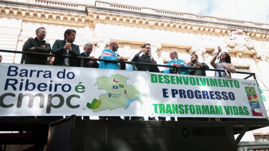 A fotografia em plano m&eacute;dio e &acirc;ngulo contrapicado (de baixo para cima) registra um ato p&uacute;blico com autoridades e lideran&ccedil;as pol&iacute;ticas sobre a estrutura de um caminh&atilde;o de som (trio el&eacute;trico). O t&iacute;tulo do arquivo contextualiza o evento como o refor&ccedil;o do Governo do Estado &agrave; regularidade do processo de licenciamento da empresa CMPC em Barra do Ribeiro.

O Grupo e a A&ccedil;&atilde;o
Discurso Central: Pr&oacute;ximo ao centro da estrutura, um homem com barba usa um microfone e gesticula com a m&atilde;o direita enquanto fala ao p&uacute;blico. Ele veste um colete escuro sobre uma camisa social.

Autoridades e Apoiadores: Ao redor do orador, cerca de 10 pessoas acompanham o pronunciamento apoiadas no parapeito met&aacute;lico do ve&iacute;culo. Duas delas (um homem &agrave; esquerda do orador e uma mulher &agrave; direita) usam bandeiras amarradas sobre os ombros nos tons de azul, branco e vermelho. Na extrema direita, uma mulher levanta o bra&ccedil;o segurando um smartphone para registrar o momento. Os participantes vestem trajes casuais de inverno, como jaquetas estofadas e casacos.

Banners e Identifica&ccedil;&atilde;o Visual
Na lateral do caminh&atilde;o de som, h&aacute; duas grandes faixas brancas horizontais com mensagens de apoio ao projeto:

Faixa da Esquerda: Exibe a frase em letras escuras e verdes: "Barra do Ribeiro &eacute; cmpc". Ao centro, h&aacute; uma ilustra&ccedil;&atilde;o do mapa do munic&iacute;pio de Barra do Ribeiro com um marcador de localiza&ccedil;&atilde;o azul e pequenas bandeiras (incluindo as do Chile e do Brasil) desenhadas sobre o mapa.

Faixa da Direita: Traz os dizeres em letras mai&uacute;sculas verdes e azuis: "DESENVOLVIMENTO E PROGRESSO TRANSFORMAM VIDAS". Na lateral direita desta faixa, aparecem verticalmente os logotipos da CMPC e pequenas bandeiras estilizadas (Brasil, Rio Grande do Sul e Chile).

O Ambiente
Cen&aacute;rio Urbano: O caminh&atilde;o de som est&aacute; posicionado em frente a um edif&iacute;cio hist&oacute;rico imponente de arquitetura cl&aacute;ssica. A fachada exibe detalhes ornamentados em relevo na pedra, colunas trabalhadas e janelas altas.

Ilumina&ccedil;&atilde;o: A foto foi tirada em um ambiente externo com luz natural diurna. O c&eacute;u aparece parcialmente vis&iacute;vel na parte superior, claro e com poucas nuvens.