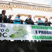 A fotografia em plano m&eacute;dio e &acirc;ngulo contrapicado (de baixo para cima) registra um ato p&uacute;blico com autoridades e lideran&ccedil;as pol&iacute;ticas sobre a estrutura de um caminh&atilde;o de som (trio el&eacute;trico). O t&iacute;tulo do arquivo contextualiza o evento como o refor&ccedil;o do Governo do Estado &agrave; regularidade do processo de licenciamento da empresa CMPC em Barra do Ribeiro.

O Grupo e a A&ccedil;&atilde;o
Discurso Central: Pr&oacute;ximo ao centro da estrutura, um homem com barba usa um microfone e gesticula com a m&atilde;o direita enquanto fala ao p&uacute;blico. Ele veste um colete escuro sobre uma camisa social.

Autoridades e Apoiadores: Ao redor do orador, cerca de 10 pessoas acompanham o pronunciamento apoiadas no parapeito met&aacute;lico do ve&iacute;culo. Duas delas (um homem &agrave; esquerda do orador e uma mulher &agrave; direita) usam bandeiras amarradas sobre os ombros nos tons de azul, branco e vermelho. Na extrema direita, uma mulher levanta o bra&ccedil;o segurando um smartphone para registrar o momento. Os participantes vestem trajes casuais de inverno, como jaquetas estofadas e casacos.

Banners e Identifica&ccedil;&atilde;o Visual
Na lateral do caminh&atilde;o de som, h&aacute; duas grandes faixas brancas horizontais com mensagens de apoio ao projeto:

Faixa da Esquerda: Exibe a frase em letras escuras e verdes: "Barra do Ribeiro &eacute; cmpc". Ao centro, h&aacute; uma ilustra&ccedil;&atilde;o do mapa do munic&iacute;pio de Barra do Ribeiro com um marcador de localiza&ccedil;&atilde;o azul e pequenas bandeiras (incluindo as do Chile e do Brasil) desenhadas sobre o mapa.

Faixa da Direita: Traz os dizeres em letras mai&uacute;sculas verdes e azuis: "DESENVOLVIMENTO E PROGRESSO TRANSFORMAM VIDAS". Na lateral direita desta faixa, aparecem verticalmente os logotipos da CMPC e pequenas bandeiras estilizadas (Brasil, Rio Grande do Sul e Chile).

O Ambiente
Cen&aacute;rio Urbano: O caminh&atilde;o de som est&aacute; posicionado em frente a um edif&iacute;cio hist&oacute;rico imponente de arquitetura cl&aacute;ssica. A fachada exibe detalhes ornamentados em relevo na pedra, colunas trabalhadas e janelas altas.

Ilumina&ccedil;&atilde;o: A foto foi tirada em um ambiente externo com luz natural diurna. O c&eacute;u aparece parcialmente vis&iacute;vel na parte superior, claro e com poucas nuvens.