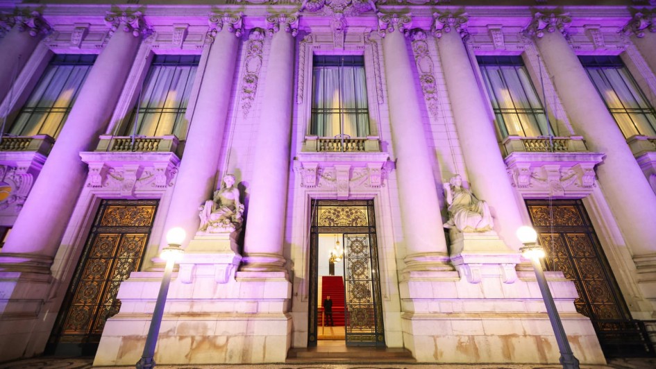 A fotografia em plano geral e &acirc;ngulo contrapicado (de baixo para cima) registra a imponente fachada do Pal&aacute;cio Piratini iluminada de forma tem&aacute;tica. O t&iacute;tulo do arquivo associa a a&ccedil;&atilde;o &agrave; celebra&ccedil;&atilde;o dos **25 anos do Teste do Pezinho** e ao refor&ccedil;o da import&acirc;ncia do diagn&oacute;stico precoce no SUS.

### **Arquitetura e Ilumina&ccedil;&atilde;o**

* **A Ilumina&ccedil;&atilde;o Tem&aacute;tica:** Toda a imponente estrutura de pedra da fachada hist&oacute;rica est&aacute; banhada por uma ilumina&ccedil;&atilde;o c&ecirc;nica noturna em tons de lil&aacute;s e roxo. A luz colore as grandes colunas, os relevos ornamentados e as sacadas do edif&iacute;cio.
* **Elementos da Fachada:** A composi&ccedil;&atilde;o destaca a simetria cl&aacute;ssica do pal&aacute;cio, com altas colunas cil&iacute;ndricas que sustentam a parte superior da estrutura, janelas verticais compridas protegidas por cortinas claras no andar superior, e portas de ferro trabalhado no t&eacute;rreo. Um grande bras&atilde;o esculpido em pedra encima a entrada principal.
* **Est&aacute;tuas e Adornos:** Ladeando o portal central, h&aacute; duas grandes esculturas de m&aacute;rmore claro representando figuras humanas sentadas sobre pedestais integrados &agrave; arquitetura.

### **Entrada Principal e Interior**

* **O Portal Central:** A grande porta principal de ferro e vidro est&aacute; parcialmente aberta. Atrav&eacute;s dela, &eacute; poss&iacute;vel vislumbrar o interior iluminado do pal&aacute;cio, revelando um lustre cl&aacute;ssico aceso e uma escadaria interna imponente revestida com um tapete vermelho vivo.
* **Seguran&ccedil;a:** Uma silhueta escura de um seguran&ccedil;a ou guarda em p&eacute; &eacute; vis&iacute;vel no interior da entrada, logo &agrave; frente dos primeiros degraus da escada.

### **Primeiro Plano**

* **Ilumina&ccedil;&atilde;o P&uacute;blica:** No n&iacute;vel da cal&ccedil;ada, em frente ao pr&eacute;dio, dois postes de ilumina&ccedil;&atilde;o de estilo antigo (lampi&otilde;es) com globos esf&eacute;ricos brancos acesos est&atilde;o posicionados um de cada lado, projetando uma luz amarelada e quente na base da fachada.
