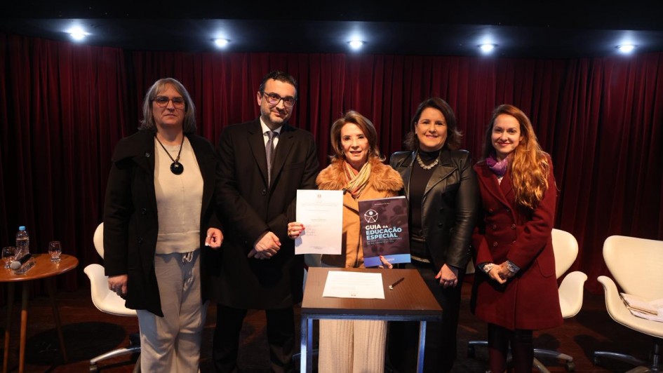 Esta &eacute; uma descri&ccedil;&atilde;o da imagem para fins de acessibilidade:

A fotografia registra um ato institucional realizado em um palco, com foco na apresenta&ccedil;&atilde;o de um documento norteador, identificado pelo t&iacute;tulo do arquivo como uma orienta&ccedil;&atilde;o para escolas e profissionais refor&ccedil;ando o compromisso com a aprendizagem.

### **Os Participantes e a A&ccedil;&atilde;o Central**

* **O Grupo:** Cinco pessoas (quatro mulheres e um homem) est&atilde;o posicionadas lado a lado em p&eacute;, voltadas para a frente e sorrindo para a fotografia.
* **A&ccedil;&atilde;o Principal:** Centralizados no grupo, o homem e a mulher ao seu lado seguram juntos uma folha de papel branca com um documento oficial impresso. A mulher tamb&eacute;m segura uma cartilha ou livro com capa escura e detalhes em azul, onde se l&ecirc; em letras mai&uacute;sculas brancas: **"GUIA DA EDUCA&Ccedil;&Atilde;O ESPECIAL"**.
* **Composi&ccedil;&atilde;o (da esquerda para a direita):**
* Uma mulher de &oacute;culos, vestindo um casaco preto longo aberto sobre uma blusa clara e cal&ccedil;a cinza-clara, usando um colar com pingente circular escuro.
* Um homem de &oacute;culos, trajando terno escuro, camisa social e gravata.
* Uma mulher com casaco marrom claro com gola de pele estilizada e cachecol colorido.
* Uma mulher de casaco ou jaqueta de material sint&eacute;tico preto brilhante sobre blusa escura e usando um colar met&aacute;lico.
* Uma mulher de cabelos longos e claros, vestindo um casaco sobretudo vermelho-escuro (vinho) e um len&ccedil;o lil&aacute;s amarrado ao pesco&ccedil;o.



### **O Ambiente e Elementos do Palco**

* **Mobili&aacute;rio:** &Agrave; frente do grupo, h&aacute; uma pequena mesa auxiliar quadrada de madeira escura com uma folha de papel e uma caneta sobre ela, indicando um ato de assinatura. Atr&aacute;s das pessoas, veem-se cadeiras de escrit&oacute;rio estofadas na cor branca com bases girat&oacute;rias cromadas. No canto esquerdo, h&aacute; uma pequena mesa redonda de madeira que apoia copos e uma garrafa de &aacute;gua.
* **Cen&aacute;rio:** O fundo do palco &eacute; composto por uma grande cortina de tecido plissado na cor vermelha escura ou bord&ocirc;. O teto rebaixado preto possui uma fileira de spots de luz embutidos acesos que iluminam o grupo diretamente. O piso &eacute; de t&aacute;buas de madeira escura polida.