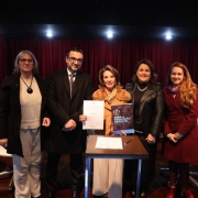 Esta &eacute; uma descri&ccedil;&atilde;o da imagem para fins de acessibilidade:

A fotografia registra um ato institucional realizado em um palco, com foco na apresenta&ccedil;&atilde;o de um documento norteador, identificado pelo t&iacute;tulo do arquivo como uma orienta&ccedil;&atilde;o para escolas e profissionais refor&ccedil;ando o compromisso com a aprendizagem.

### **Os Participantes e a A&ccedil;&atilde;o Central**

* **O Grupo:** Cinco pessoas (quatro mulheres e um homem) est&atilde;o posicionadas lado a lado em p&eacute;, voltadas para a frente e sorrindo para a fotografia.
* **A&ccedil;&atilde;o Principal:** Centralizados no grupo, o homem e a mulher ao seu lado seguram juntos uma folha de papel branca com um documento oficial impresso. A mulher tamb&eacute;m segura uma cartilha ou livro com capa escura e detalhes em azul, onde se l&ecirc; em letras mai&uacute;sculas brancas: **"GUIA DA EDUCA&Ccedil;&Atilde;O ESPECIAL"**.
* **Composi&ccedil;&atilde;o (da esquerda para a direita):**
* Uma mulher de &oacute;culos, vestindo um casaco preto longo aberto sobre uma blusa clara e cal&ccedil;a cinza-clara, usando um colar com pingente circular escuro.
* Um homem de &oacute;culos, trajando terno escuro, camisa social e gravata.
* Uma mulher com casaco marrom claro com gola de pele estilizada e cachecol colorido.
* Uma mulher de casaco ou jaqueta de material sint&eacute;tico preto brilhante sobre blusa escura e usando um colar met&aacute;lico.
* Uma mulher de cabelos longos e claros, vestindo um casaco sobretudo vermelho-escuro (vinho) e um len&ccedil;o lil&aacute;s amarrado ao pesco&ccedil;o.



### **O Ambiente e Elementos do Palco**

* **Mobili&aacute;rio:** &Agrave; frente do grupo, h&aacute; uma pequena mesa auxiliar quadrada de madeira escura com uma folha de papel e uma caneta sobre ela, indicando um ato de assinatura. Atr&aacute;s das pessoas, veem-se cadeiras de escrit&oacute;rio estofadas na cor branca com bases girat&oacute;rias cromadas. No canto esquerdo, h&aacute; uma pequena mesa redonda de madeira que apoia copos e uma garrafa de &aacute;gua.
* **Cen&aacute;rio:** O fundo do palco &eacute; composto por uma grande cortina de tecido plissado na cor vermelha escura ou bord&ocirc;. O teto rebaixado preto possui uma fileira de spots de luz embutidos acesos que iluminam o grupo diretamente. O piso &eacute; de t&aacute;buas de madeira escura polida.