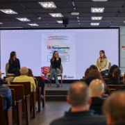 A fotografia em plano geral e &acirc;ngulo frontal registra a realiza&ccedil;&atilde;o de um painel de apresenta&ccedil;&atilde;o em um audit&oacute;rio, identificado pelo t&iacute;tulo do arquivo como o lan&ccedil;amento da **Edi&ccedil;&atilde;o especial da revista Estudos de Planejamento apresenta papel das Rela&ccedil;&otilde;es Internacionais no planejamento p&uacute;blico**.

### **O Palco e as Integrantes**

* **A&ccedil;&atilde;o Central:** No centro do palco, uma mulher de &oacute;culos, blusa preta de gola alta e cal&ccedil;a jeans clara est&aacute; sentada em uma cadeira, segurando um microfone e falando ao p&uacute;blico.
* **Demais Painelistas:** Ela &eacute; ladeada por outras duas mulheres sentadas. &Agrave; esquerda, uma mulher veste um conjunto escuro (blazer e cal&ccedil;a); &agrave; direita, outra mulher usa um casaco de tric&ocirc; amarelo-claro com um len&ccedil;o vermelho estampado amarrado ao pesco&ccedil;o e um crach&aacute; institucional.

### **O Painel Digital de Fundo**

* **Proje&ccedil;&atilde;o:** Atr&aacute;s das painelistas, uma grande tela digital estende-se por quase toda a parede do fundo, exibindo a capa da publica&ccedil;&atilde;o t&eacute;cnica em formato digital.
* **Elementos da Capa:** No slide projetado, l&ecirc;-se o t&iacute;tulo em destaque: **"Estudos de Planejamento - Edi&ccedil;&atilde;o Especial de Rela&ccedil;&otilde;es Internacionais"**. O documento traz grafado na parte superior as inst&acirc;ncias institucionais: "Governo do Estado do Rio Grande do Sul / Secretaria de Planejamento, Governan&ccedil;a e Gest&atilde;o / Subsecretaria de Planejamento / Departamento de Planejamento Governamental". Na parte inferior da proje&ccedil;&atilde;o, veem-se ilustra&ccedil;&otilde;es estilizadas de um conjunto de bandeiras de diferentes pa&iacute;ses e o logotipo oficial do governo do estado.

### **O Ambiente e a Plateia**

* **A Plateia:** Em primeiro plano, v&aacute;rias fileiras de poltronas de madeira com encosto castanho est&atilde;o preenchidas por espectadores assistindo &agrave; apresenta&ccedil;&atilde;o, vistos predominantemente de costas.
* **Infraestrutura do Local:** O audit&oacute;rio possui teto rebaixado de placas pretas quadriculadas, equipado com diversas lumin&aacute;rias de LED quadradas embutidas e acesas. &Agrave; direita da tela, h&aacute; uma mureta de vidro com uma placa de sinaliza&ccedil;&atilde;o vertical vermelha e branca onde se l&ecirc; em letras mai&uacute;sculas: **"AVISO / N&Atilde;O ENCOSTE NO TEL&Atilde;O"**.