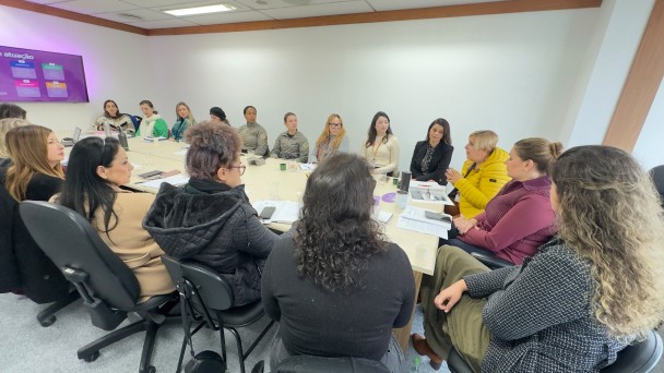 Sala de reuni&atilde;o com mesa retangular clara cercada por v&aacute;rias pessoas sentadas, com cadernos, celulares e copos de &aacute;gua distribu&iacute;dos sobre a superf&iacute;cie.