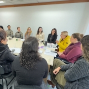 Sala de reuni&atilde;o com mesa retangular clara cercada por v&aacute;rias pessoas sentadas, com cadernos, celulares e copos de &aacute;gua distribu&iacute;dos sobre a superf&iacute;cie.