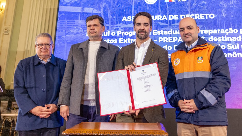 A fotografia em plano m&eacute;dio e &acirc;ngulo frontal registra um ato oficial com autoridades estaduais, identificado pelo nome do arquivo como a assinatura de contrato pelo **Governo do Estado assina contrato para constru&ccedil;&atilde;o do Centro de Log&iacute;stica Humanit&aacute;ria.jpg**.

### **Os Participantes e a A&ccedil;&atilde;o**

* **O Grupo:** Quatro homens est&atilde;o posicionados em p&eacute;, lado a lado, voltados para a frente em postura formal.
* **A&ccedil;&atilde;o Principal:** O homem posicionado ao centro-direito segura e exibe uma pasta oficial aberta com bordas vermelhas, contendo um documento assinado em folha branca. Ele segura uma caneta preta com a m&atilde;o direita.
* **Composi&ccedil;&atilde;o (da esquerda para a direita):**
* O primeiro homem usa &oacute;culos de arma&ccedil;&atilde;o escura, camisa clara e uma jaqueta azul-marinho abotoada at&eacute; o peito. Ele mant&eacute;m as m&atilde;os unidas &agrave; frente do corpo.
* O segundo homem veste um su&eacute;ter de gola alta branco sob um casaco sobretudo cinza aberto e cal&ccedil;a escura.
* O terceiro homem (o que segura o documento) possui barba aparada e veste blazer marrom sobre camisa social branca sem gravata.
* O quarto homem, posicionado na extrema direita, veste um uniforme oficial operacional composto por uma jaqueta azul-escuro com uma ampla faixa horizontal amarela ou laranja no peito e ombros, contendo dois bras&otilde;es institucionais bordados (incluindo o da Defesa Civil). Ele usa cal&ccedil;a bege e est&aacute; com as m&atilde;os unidas &agrave; frente.



### **O Painel de Fundo e o Ambiente**

* **Tel&atilde;o Digital:** Atr&aacute;s do grupo, uma grande tela com fundo azul exibe textos em letras mai&uacute;sculas brancas. L&ecirc;-se em destaque na linha superior: **"ASSINATURA DE DECRETO"**, seguido por linhas textuais parcialmente cobertas pelas autoridades, onde se identificam termos como "institui o Programa Estadual de Prepara&ccedil;&atilde;o para Eventos Extremos - PREPARAR RS". No topo superior direito da tela, constam os logotipos oficiais da Defesa Civil (Casa Militar) e do Governo do Estado do Rio Grande do Sul.
* **Mobili&aacute;rio e Cen&aacute;rio:** Em primeiro plano, logo abaixo do documento exibido, v&ecirc;-se a parte superior de uma mesa auxiliar de madeira com bordas trabalhadas em relevo. No canto esquerdo, ao fundo, nota-se uma coluna de arquitetura cl&aacute;ssica em tom bege claro pertencente ao sal&atilde;o do evento.