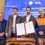 A fotografia em plano m&eacute;dio e &acirc;ngulo frontal registra um ato oficial com autoridades estaduais, identificado pelo nome do arquivo como a assinatura de contrato pelo **Governo do Estado assina contrato para constru&ccedil;&atilde;o do Centro de Log&iacute;stica Humanit&aacute;ria.jpg**.

### **Os Participantes e a A&ccedil;&atilde;o**

* **O Grupo:** Quatro homens est&atilde;o posicionados em p&eacute;, lado a lado, voltados para a frente em postura formal.
* **A&ccedil;&atilde;o Principal:** O homem posicionado ao centro-direito segura e exibe uma pasta oficial aberta com bordas vermelhas, contendo um documento assinado em folha branca. Ele segura uma caneta preta com a m&atilde;o direita.
* **Composi&ccedil;&atilde;o (da esquerda para a direita):**
* O primeiro homem usa &oacute;culos de arma&ccedil;&atilde;o escura, camisa clara e uma jaqueta azul-marinho abotoada at&eacute; o peito. Ele mant&eacute;m as m&atilde;os unidas &agrave; frente do corpo.
* O segundo homem veste um su&eacute;ter de gola alta branco sob um casaco sobretudo cinza aberto e cal&ccedil;a escura.
* O terceiro homem (o que segura o documento) possui barba aparada e veste blazer marrom sobre camisa social branca sem gravata.
* O quarto homem, posicionado na extrema direita, veste um uniforme oficial operacional composto por uma jaqueta azul-escuro com uma ampla faixa horizontal amarela ou laranja no peito e ombros, contendo dois bras&otilde;es institucionais bordados (incluindo o da Defesa Civil). Ele usa cal&ccedil;a bege e est&aacute; com as m&atilde;os unidas &agrave; frente.



### **O Painel de Fundo e o Ambiente**

* **Tel&atilde;o Digital:** Atr&aacute;s do grupo, uma grande tela com fundo azul exibe textos em letras mai&uacute;sculas brancas. L&ecirc;-se em destaque na linha superior: **"ASSINATURA DE DECRETO"**, seguido por linhas textuais parcialmente cobertas pelas autoridades, onde se identificam termos como "institui o Programa Estadual de Prepara&ccedil;&atilde;o para Eventos Extremos - PREPARAR RS". No topo superior direito da tela, constam os logotipos oficiais da Defesa Civil (Casa Militar) e do Governo do Estado do Rio Grande do Sul.
* **Mobili&aacute;rio e Cen&aacute;rio:** Em primeiro plano, logo abaixo do documento exibido, v&ecirc;-se a parte superior de uma mesa auxiliar de madeira com bordas trabalhadas em relevo. No canto esquerdo, ao fundo, nota-se uma coluna de arquitetura cl&aacute;ssica em tom bege claro pertencente ao sal&atilde;o do evento.
