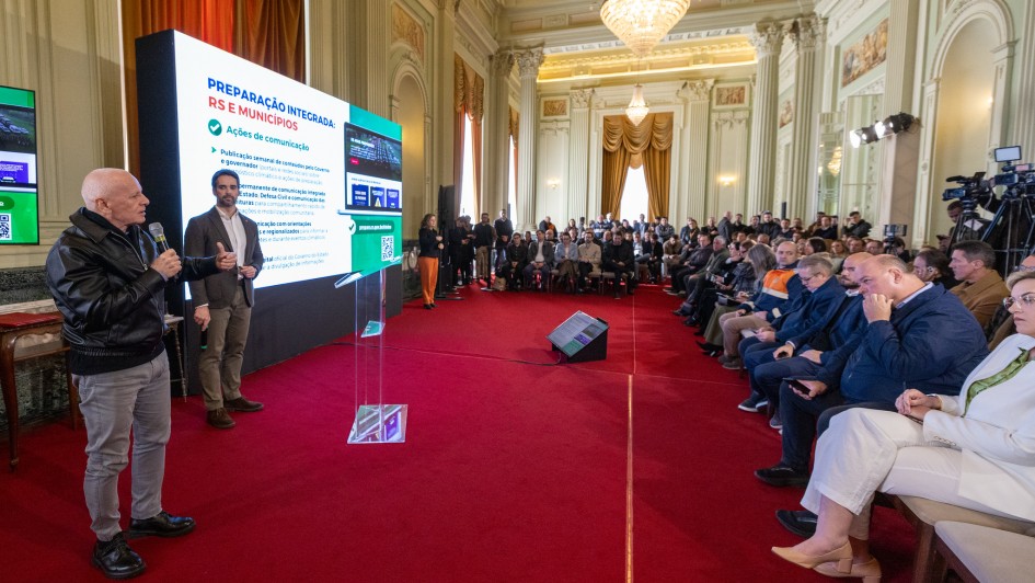 Ambiente interno de evento institucional, onde uma pessoa em primeiro plano segura um microfone e fala diante de um p&uacute;lpito transparente.