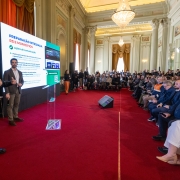 Ambiente interno de evento institucional, onde uma pessoa em primeiro plano segura um microfone e fala diante de um p&uacute;lpito transparente.