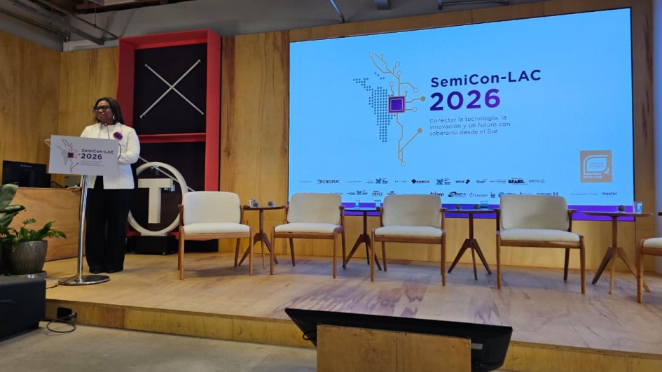 A fotografia em plano geral e &acirc;ngulo frontal registra a abertura de um evento institucional e tecnol&oacute;gico em um palco, identificado pelo t&iacute;tulo do arquivo como **Abertura do SemiCon-LAC 2026 posiciona o RS como hub estrat&eacute;gico de semicondutores na Am&eacute;rica Latina.jpeg**.

### **A Palestrante**

* **A&ccedil;&atilde;o e Traje:** No lado esquerdo do palco, uma mulher negra usa &oacute;culos de grau e est&aacute; em p&eacute; atr&aacute;s de um p&uacute;lpito branco. Ela fala ao p&uacute;blico com o corpo voltado ligeiramente para a direita.
* **Vestu&aacute;rio:** Ela veste um blazer branco estruturado com uma flor roxa ou broche fixado na lapela esquerda, sobre uma blusa clara, e cal&ccedil;a comprida preta de corte reto.

### **O P&uacute;lpito e o Cen&aacute;rio do Palco**

* **P&uacute;lpito:** A face frontal do p&uacute;lpito traz a identidade visual do evento com a inscri&ccedil;&atilde;o **"SemiCon-LAC 2026"**.
* **Estrutura de Fundo:** Atr&aacute;s da palestrante, h&aacute; um painel decorativo vertical preto com uma moldura vermelha volumosa. No topo desse painel, duas barras brancas cruzadas formam a letra "X". Logo abaixo, destaca-se um letreiro luminoso circular branco contendo a letra **"T"** em seu interior.
* **Mobili&aacute;rio:** Ao longo do palco revestido de madeira clara, h&aacute; uma fileira de quatro poltronas estofadas na cor branca ou creme com bra&ccedil;os e pernas de madeira clara. Entre as poltronas, est&atilde;o dispostas pequenas mesas de apoio redondas de tr&ecirc;s p&eacute;s, tamb&eacute;m de madeira, sustentando copos de &aacute;gua.

### **O Painel Digital e Conte&uacute;do**

* **O Tel&atilde;o:** &Agrave; direita, um grande painel digital de LED exibe o slide principal de abertura do evento com fundo branco.
* **Inscri&ccedil;&otilde;es e Gr&aacute;ficos:** No centro do slide, destaca-se o t&iacute;tulo em letras escuras e roxas: **"SemiCon-LAC 2026"**. Abaixo do t&iacute;tulo, l&ecirc;-se o slogan em espanhol: *"Conectar la tecnolog&iacute;a, la innovaci&oacute;n y un futuro con soberan&iacute;a desde el Sur"*. &Agrave; esquerda do texto, h&aacute; uma ilustra&ccedil;&atilde;o digital pontilhada que simula o mapa da Am&eacute;rica Latina interconectado por linhas que remetem a circuitos integrados ou semicondutores.
* **Logotipos de Apoio:** Na borda inferior do tel&atilde;o, h&aacute; uma linha horizontal contendo diversos logotipos pequenos de institui&ccedil;&otilde;es parceiras e realizadoras, sendo poss&iacute;vel identificar o nome **"TECNOPUC"** na extremidade esquerda. No canto inferior direito da tela, h&aacute; um selo quadrado laranja.

### **O Ambiente e Ilumina&ccedil;&atilde;o**

* **Infraestrutura:** O palco possui paredes laterais com fechamento em pain&eacute;is de madeira clara. O teto do local &eacute; industrial e exposto, revelando vigas de concreto, tubula&ccedil;&otilde;es de metal e dutos de ar-condicionado prateados.
* **Primeiro Plano:** Em primeiro plano, abaixo do n&iacute;vel do palco, o ch&atilde;o &eacute; de concreto cinza polido e h&aacute; um monitor de retorno preto inclinado e voltado para as poltronas, sustentado por uma base de madeira. No canto esquerdo inferior, avista-se um vaso escuro com folhagens verdes. A ilumina&ccedil;&atilde;o &eacute; difusa e predominantemente direcionada para o palco e para o tel&atilde;o.