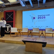 A fotografia em plano geral e &acirc;ngulo frontal registra a abertura de um evento institucional e tecnol&oacute;gico em um palco, identificado pelo t&iacute;tulo do arquivo como **Abertura do SemiCon-LAC 2026 posiciona o RS como hub estrat&eacute;gico de semicondutores na Am&eacute;rica Latina.jpeg**.

### **A Palestrante**

* **A&ccedil;&atilde;o e Traje:** No lado esquerdo do palco, uma mulher negra usa &oacute;culos de grau e est&aacute; em p&eacute; atr&aacute;s de um p&uacute;lpito branco. Ela fala ao p&uacute;blico com o corpo voltado ligeiramente para a direita.
* **Vestu&aacute;rio:** Ela veste um blazer branco estruturado com uma flor roxa ou broche fixado na lapela esquerda, sobre uma blusa clara, e cal&ccedil;a comprida preta de corte reto.

### **O P&uacute;lpito e o Cen&aacute;rio do Palco**

* **P&uacute;lpito:** A face frontal do p&uacute;lpito traz a identidade visual do evento com a inscri&ccedil;&atilde;o **"SemiCon-LAC 2026"**.
* **Estrutura de Fundo:** Atr&aacute;s da palestrante, h&aacute; um painel decorativo vertical preto com uma moldura vermelha volumosa. No topo desse painel, duas barras brancas cruzadas formam a letra "X". Logo abaixo, destaca-se um letreiro luminoso circular branco contendo a letra **"T"** em seu interior.
* **Mobili&aacute;rio:** Ao longo do palco revestido de madeira clara, h&aacute; uma fileira de quatro poltronas estofadas na cor branca ou creme com bra&ccedil;os e pernas de madeira clara. Entre as poltronas, est&atilde;o dispostas pequenas mesas de apoio redondas de tr&ecirc;s p&eacute;s, tamb&eacute;m de madeira, sustentando copos de &aacute;gua.

### **O Painel Digital e Conte&uacute;do**

* **O Tel&atilde;o:** &Agrave; direita, um grande painel digital de LED exibe o slide principal de abertura do evento com fundo branco.
* **Inscri&ccedil;&otilde;es e Gr&aacute;ficos:** No centro do slide, destaca-se o t&iacute;tulo em letras escuras e roxas: **"SemiCon-LAC 2026"**. Abaixo do t&iacute;tulo, l&ecirc;-se o slogan em espanhol: *"Conectar la tecnolog&iacute;a, la innovaci&oacute;n y un futuro con soberan&iacute;a desde el Sur"*. &Agrave; esquerda do texto, h&aacute; uma ilustra&ccedil;&atilde;o digital pontilhada que simula o mapa da Am&eacute;rica Latina interconectado por linhas que remetem a circuitos integrados ou semicondutores.
* **Logotipos de Apoio:** Na borda inferior do tel&atilde;o, h&aacute; uma linha horizontal contendo diversos logotipos pequenos de institui&ccedil;&otilde;es parceiras e realizadoras, sendo poss&iacute;vel identificar o nome **"TECNOPUC"** na extremidade esquerda. No canto inferior direito da tela, h&aacute; um selo quadrado laranja.

### **O Ambiente e Ilumina&ccedil;&atilde;o**

* **Infraestrutura:** O palco possui paredes laterais com fechamento em pain&eacute;is de madeira clara. O teto do local &eacute; industrial e exposto, revelando vigas de concreto, tubula&ccedil;&otilde;es de metal e dutos de ar-condicionado prateados.
* **Primeiro Plano:** Em primeiro plano, abaixo do n&iacute;vel do palco, o ch&atilde;o &eacute; de concreto cinza polido e h&aacute; um monitor de retorno preto inclinado e voltado para as poltronas, sustentado por uma base de madeira. No canto esquerdo inferior, avista-se um vaso escuro com folhagens verdes. A ilumina&ccedil;&atilde;o &eacute; difusa e predominantemente direcionada para o palco e para o tel&atilde;o.