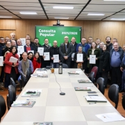 Foto mostra o secret&aacute;rio, os prefeitos e assessores posando para foto em uma sala de reuni&otilde;es. Ao fundo, uma tela verde mostra o nome Consulta Popular .
