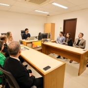 A imagem mostra uma sala de reuni&otilde;es, com pessoas sentadas ao redor de mesas de escrit&oacute;rio. 