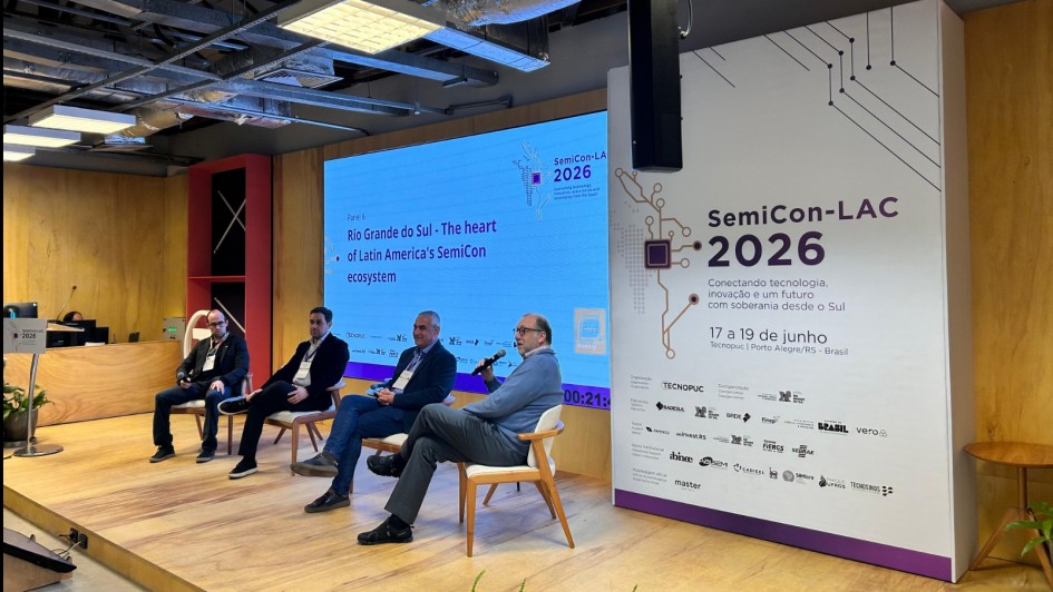 Foto mostra palestrantes debatendo no palco do evento. Eles est&atilde;o sentados em poltronas, com um tel&atilde;o ao fundo. Um painel branco e lil&aacute;s mostra a logomarca do evento. 