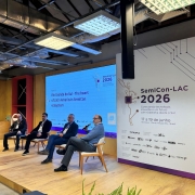 Foto mostra palestrantes debatendo no palco do evento. Eles est&atilde;o sentados em poltronas, com um tel&atilde;o ao fundo. Um painel branco e lil&aacute;s mostra a logomarca do evento. 
