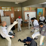 Foto mostra uma aula de capoeira dentro de uma sala de aula. O mestre faz um movimento que &eacute; imitado pelos demais participantes. 