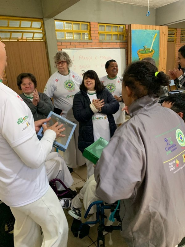 Foto mostra professor e alunos tocando instrumentos musicais produzidos por eles, no formato de uma caixa retangular. Outros alunos acompanham com palmas.