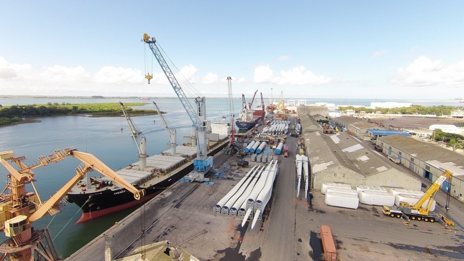 A fotografia a&eacute;rea apresenta uma vista panor&acirc;mica de um porto comercial em plena atividade. O cen&aacute;rio &eacute; dominado por grandes infraestruturas log&iacute;sticas, navios de carga e componentes industriais sob um c&eacute;u claro com algumas nuvens esparsas. 