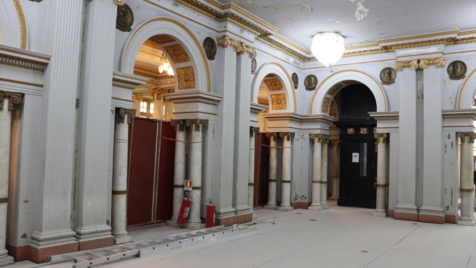 Esta &eacute; uma descri&ccedil;&atilde;o da imagem para fins de acessibilidade:

A fotografia em plano geral e &acirc;ngulo frontal registra o sal&atilde;o interno de um pal&aacute;cio ou edif&iacute;cio hist&oacute;rico passando por obras de restaura&ccedil;&atilde;o ou reforma estrutural. O t&iacute;tulo do arquivo indica a a&ccedil;&atilde;o com a inscri&ccedil;&atilde;o **"Com investimento superior a R$ 12,6 milh&otilde;es.jpg"**.

### **Arquitetura Cl&aacute;ssica e Ornamentos**

* **Colunas e Arcos:** O amplo sal&atilde;o possui uma sucess&atilde;o de altas colunas decorativas acopladas &agrave;s paredes (pilastras) em tom cinza-claro, intercaladas por grandes arcos de passagem abertos. Abaixo de cada arco principal, h&aacute; colunas menores e cil&iacute;ndricas feitas de m&aacute;rmore claro com veios acinzentados e capit&eacute;is detalhados de estilo cl&aacute;ssico.
* **Detalhes em Ouro:** A parte superior das paredes exibe frisos e molduras horizontais ornamentadas com pintura dourada envelhecida. O interior dos arcos centrais tamb&eacute;m possui relevos em formato de flores decorados em dourado.
* **Medalh&otilde;es Hist&oacute;ricos:** Fixados nas paredes logo acima dos capit&eacute;is das pilastras, veem-se medalh&otilde;es circulares de bronze ou metal escuro exibindo rostos em relevo (ef&iacute;gies) de figuras hist&oacute;ricas de perfil.

### **Elementos da Obra e Prote&ccedil;&atilde;o**

* **Piso Protegido:** Todo o ch&atilde;o do sal&atilde;o est&aacute; totalmente coberto e protegido por placas retangulares cont&iacute;nuas de material protetor claro (como compensado, MDF ou papel&atilde;o estruturado na cor bege), ocultando o piso original.
* **Isolamentos:** Os v&atilde;os dos arcos localizados no lado esquerdo est&atilde;o temporariamente bloqueados por tapumes verticais de madeira na cor vermelha escura ou marrom, isolando as &aacute;reas internas cont&iacute;guas.
* **Equipamentos:** Uma escada de alum&iacute;nio extens&iacute;vel est&aacute; deitada horizontalmente sobre o piso protegido, paralela &agrave; base das colunas da esquerda. Pr&oacute;ximo a ela, encostados na base de uma pilastra, veem-se dois extintores de inc&ecirc;ndio cil&iacute;ndricos vermelhos e uma placa de sinaliza&ccedil;&atilde;o.

### **Teto e Ilumina&ccedil;&atilde;o**

* **Estado de Conserva&ccedil;&atilde;o:** O teto de gesso ou alvenaria clara apresenta evidentes sinais de desgaste hist&oacute;rico, com descascamentos, manchas e infiltra&ccedil;&otilde;es na pintura que revelam camadas subjacentes amareladas, evidenciando a necessidade de restauro.
* **Lumin&aacute;rias:** Grandes globos de ilumina&ccedil;&atilde;o suspensos por correntes est&atilde;o protegidos por sacos ou pl&aacute;sticos brancos amarrados na base, evitando o ac&uacute;mulo de poeira decorrente das obras. Atrav&eacute;s de um dos v&atilde;os ao fundo, &eacute; poss&iacute;vel vislumbrar o brilho amarelado de um lustre aceso em outra sala.