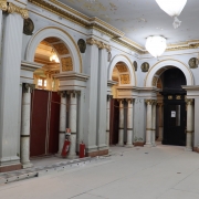 Esta &eacute; uma descri&ccedil;&atilde;o da imagem para fins de acessibilidade:

A fotografia em plano geral e &acirc;ngulo frontal registra o sal&atilde;o interno de um pal&aacute;cio ou edif&iacute;cio hist&oacute;rico passando por obras de restaura&ccedil;&atilde;o ou reforma estrutural. O t&iacute;tulo do arquivo indica a a&ccedil;&atilde;o com a inscri&ccedil;&atilde;o **"Com investimento superior a R$ 12,6 milh&otilde;es.jpg"**.

### **Arquitetura Cl&aacute;ssica e Ornamentos**

* **Colunas e Arcos:** O amplo sal&atilde;o possui uma sucess&atilde;o de altas colunas decorativas acopladas &agrave;s paredes (pilastras) em tom cinza-claro, intercaladas por grandes arcos de passagem abertos. Abaixo de cada arco principal, h&aacute; colunas menores e cil&iacute;ndricas feitas de m&aacute;rmore claro com veios acinzentados e capit&eacute;is detalhados de estilo cl&aacute;ssico.
* **Detalhes em Ouro:** A parte superior das paredes exibe frisos e molduras horizontais ornamentadas com pintura dourada envelhecida. O interior dos arcos centrais tamb&eacute;m possui relevos em formato de flores decorados em dourado.
* **Medalh&otilde;es Hist&oacute;ricos:** Fixados nas paredes logo acima dos capit&eacute;is das pilastras, veem-se medalh&otilde;es circulares de bronze ou metal escuro exibindo rostos em relevo (ef&iacute;gies) de figuras hist&oacute;ricas de perfil.

### **Elementos da Obra e Prote&ccedil;&atilde;o**

* **Piso Protegido:** Todo o ch&atilde;o do sal&atilde;o est&aacute; totalmente coberto e protegido por placas retangulares cont&iacute;nuas de material protetor claro (como compensado, MDF ou papel&atilde;o estruturado na cor bege), ocultando o piso original.
* **Isolamentos:** Os v&atilde;os dos arcos localizados no lado esquerdo est&atilde;o temporariamente bloqueados por tapumes verticais de madeira na cor vermelha escura ou marrom, isolando as &aacute;reas internas cont&iacute;guas.
* **Equipamentos:** Uma escada de alum&iacute;nio extens&iacute;vel est&aacute; deitada horizontalmente sobre o piso protegido, paralela &agrave; base das colunas da esquerda. Pr&oacute;ximo a ela, encostados na base de uma pilastra, veem-se dois extintores de inc&ecirc;ndio cil&iacute;ndricos vermelhos e uma placa de sinaliza&ccedil;&atilde;o.

### **Teto e Ilumina&ccedil;&atilde;o**

* **Estado de Conserva&ccedil;&atilde;o:** O teto de gesso ou alvenaria clara apresenta evidentes sinais de desgaste hist&oacute;rico, com descascamentos, manchas e infiltra&ccedil;&otilde;es na pintura que revelam camadas subjacentes amareladas, evidenciando a necessidade de restauro.
* **Lumin&aacute;rias:** Grandes globos de ilumina&ccedil;&atilde;o suspensos por correntes est&atilde;o protegidos por sacos ou pl&aacute;sticos brancos amarrados na base, evitando o ac&uacute;mulo de poeira decorrente das obras. Atrav&eacute;s de um dos v&atilde;os ao fundo, &eacute; poss&iacute;vel vislumbrar o brilho amarelado de um lustre aceso em outra sala.