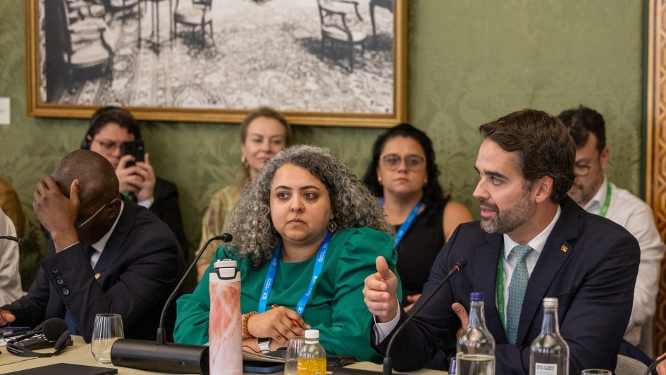 A fotografia em plano m&eacute;dio registra uma mesa de debate durante um evento oficial internacional. O t&iacute;tulo do arquivo contextualiza que o **"Estado apresenta estrat&eacute;gias do RS para transi&ccedil;&atilde;o energ&eacute;tica e resili&ecirc;ncia clim&aacute;tica em c&uacute;pula internacional em Londres.jpg"**.

### **Os Participantes da Mesa**

* **O Orador Principal:** No lado direito da imagem, em primeiro plano, o governador do estado est&aacute; sentado e voltado de perfil parcial para a esquerda enquanto fala. Ele tem barba aparada e veste um terno azul-marinho, camisa social branca, gravata texturizada em tom verde-claro ou esmeralda, e exibe um pequeno pin de bandeira oficial na lapela esquerda. Sua m&atilde;o direita est&aacute; estendida &agrave; frente em um gesto explicativo. Um microfone de haste flex&iacute;vel (pesco&ccedil;o de ganso) est&aacute; posicionado &agrave; sua frente.
* **A Integrante Central:** Sentada ao centro da mesa, ao lado do orador, uma mulher com cabelos cacheados e volumosos em tons de cinza e grisalho escuta atentamente, olhando para a esquerda. Ela veste uma blusa ou vestido verde-bandeira com mangas bufantes e usa no pesco&ccedil;o uma credencial oficial com cord&atilde;o azul. &Agrave; sua frente, sobre a mesa, h&aacute; uma garrafa t&eacute;rmica cil&iacute;ndrica com estampa abstrata em tons de rosa e branco.
* **Outros Membros da Mesa:**
* &Agrave; esquerda, um homem negro veste terno escuro e camisa clara; ele est&aacute; com a cabe&ccedil;a levemente inclinada para baixo e a m&atilde;o direita apoiada sobre a testa.
* Atr&aacute;s da mesa, em segundo plano, veem-se outros participantes e espectadores sentados, incluindo uma mulher loira &agrave; esquerda, uma mulher de &oacute;culos de grau ao centro-fundo e um homem de camisa branca com fita de credencial verde &agrave; extrema direita. Um homem ao fundo &agrave; esquerda segura um smartphone na vertical, registrando a sess&atilde;o.



### **Elementos da Mesa e Cen&aacute;rio**

* **Identifica&ccedil;&atilde;o:** Na frente do governador, sobre a mesa revestida por uma toalha bege-clara, destaca-se uma placa de identifica&ccedil;&atilde;o de papel branco em formato triangular onde se l&ecirc; em letras pretas: **"Government of Rio Grande do Sul"**.
* **Objetos:** Dispostos ao longo da mesa, veem-se garrafas de vidro com &aacute;gua mineral, copos transparentes vazios, uma pequena garrafa de suco ou bebida amarelada, fones de ouvido pretos para tradu&ccedil;&atilde;o simult&acirc;nea e bases de microfones.
* **Decora&ccedil;&atilde;o do Espa&ccedil;o:** A parede ao fundo possui um elegante revestimento texturizado ou papel de parede em tom verde-oliva sutil. Fixado na parede, no canto superior esquerdo, h&aacute; um grande quadro com moldura dourada exibindo uma fotografia antiga ou gravura hist&oacute;rica em preto e branco que retrata o sal&atilde;o de um pal&aacute;cio com diversas cadeiras e mesas cl&aacute;ssicas dispostas sobre um grande tapete decorado.