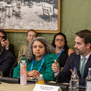 A fotografia em plano m&eacute;dio registra uma mesa de debate durante um evento oficial internacional. O t&iacute;tulo do arquivo contextualiza que o **"Estado apresenta estrat&eacute;gias do RS para transi&ccedil;&atilde;o energ&eacute;tica e resili&ecirc;ncia clim&aacute;tica em c&uacute;pula internacional em Londres.jpg"**.

### **Os Participantes da Mesa**

* **O Orador Principal:** No lado direito da imagem, em primeiro plano, o governador do estado est&aacute; sentado e voltado de perfil parcial para a esquerda enquanto fala. Ele tem barba aparada e veste um terno azul-marinho, camisa social branca, gravata texturizada em tom verde-claro ou esmeralda, e exibe um pequeno pin de bandeira oficial na lapela esquerda. Sua m&atilde;o direita est&aacute; estendida &agrave; frente em um gesto explicativo. Um microfone de haste flex&iacute;vel (pesco&ccedil;o de ganso) est&aacute; posicionado &agrave; sua frente.
* **A Integrante Central:** Sentada ao centro da mesa, ao lado do orador, uma mulher com cabelos cacheados e volumosos em tons de cinza e grisalho escuta atentamente, olhando para a esquerda. Ela veste uma blusa ou vestido verde-bandeira com mangas bufantes e usa no pesco&ccedil;o uma credencial oficial com cord&atilde;o azul. &Agrave; sua frente, sobre a mesa, h&aacute; uma garrafa t&eacute;rmica cil&iacute;ndrica com estampa abstrata em tons de rosa e branco.
* **Outros Membros da Mesa:**
* &Agrave; esquerda, um homem negro veste terno escuro e camisa clara; ele est&aacute; com a cabe&ccedil;a levemente inclinada para baixo e a m&atilde;o direita apoiada sobre a testa.
* Atr&aacute;s da mesa, em segundo plano, veem-se outros participantes e espectadores sentados, incluindo uma mulher loira &agrave; esquerda, uma mulher de &oacute;culos de grau ao centro-fundo e um homem de camisa branca com fita de credencial verde &agrave; extrema direita. Um homem ao fundo &agrave; esquerda segura um smartphone na vertical, registrando a sess&atilde;o.



### **Elementos da Mesa e Cen&aacute;rio**

* **Identifica&ccedil;&atilde;o:** Na frente do governador, sobre a mesa revestida por uma toalha bege-clara, destaca-se uma placa de identifica&ccedil;&atilde;o de papel branco em formato triangular onde se l&ecirc; em letras pretas: **"Government of Rio Grande do Sul"**.
* **Objetos:** Dispostos ao longo da mesa, veem-se garrafas de vidro com &aacute;gua mineral, copos transparentes vazios, uma pequena garrafa de suco ou bebida amarelada, fones de ouvido pretos para tradu&ccedil;&atilde;o simult&acirc;nea e bases de microfones.
* **Decora&ccedil;&atilde;o do Espa&ccedil;o:** A parede ao fundo possui um elegante revestimento texturizado ou papel de parede em tom verde-oliva sutil. Fixado na parede, no canto superior esquerdo, h&aacute; um grande quadro com moldura dourada exibindo uma fotografia antiga ou gravura hist&oacute;rica em preto e branco que retrata o sal&atilde;o de um pal&aacute;cio com diversas cadeiras e mesas cl&aacute;ssicas dispostas sobre um grande tapete decorado.