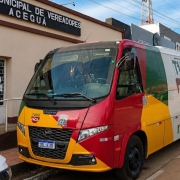 A fotografia em plano geral e &acirc;ngulo diagonal registra a unidade m&oacute;vel de atendimento ao cidad&atilde;o estacionada em uma via p&uacute;blica em frente a um pr&eacute;dio institucional. O t&iacute;tulo do arquivo identifica o contexto como a "Opera&ccedil;&atilde;o da Unidade M&oacute;vel do Tudo F&aacute;cil em Acegu&aacute; encerra com 1 mil atendimentos_.jpeg".

A Unidade M&oacute;vel
O Ve&iacute;culo: No centro do plano, destaca-se um micro-&ocirc;nibus ou van de grande porte da marca Volare, com a carroceria totalmente adesivada com a identidade visual do programa governamental.

Cores e Grafismos: O design do ve&iacute;culo combina faixas e formas geom&eacute;tricas nas cores vermelho, amarelo, verde e branco (cores que remetem &agrave; bandeira do Estado do Rio Grande do Sul).

Identidade Visual: Na lateral branca do micro-&ocirc;nibus, l&ecirc;-se em destaque o logotipo "TUDO F&Aacute;CIL" em letras verdes dentro de um formato de escudo estilizado marrom, seguido abaixo pelo endere&ccedil;o do portal oficial: "rs.gov.br". Na parte frontal amarela e vermelha, h&aacute; um adesivo menor do logotipo &agrave; esquerda e um adesivo quadrado com um c&oacute;digo QR (QR Code) centralizado acima da grade preta do radiador. A placa Mercosul do ve&iacute;culo indica a identifica&ccedil;&atilde;o "JDL1E30".

O Motorista: Atrav&eacute;s do grande para-brisa dianteiro de vidro transl&uacute;cido, &eacute; poss&iacute;vel ver o motorista ou operador no banco de condu&ccedil;&atilde;o, sorrindo e fazendo o sinal de "paz e amor" (ou "V" de vit&oacute;ria) com os dedos indicador e m&eacute;dio da m&atilde;o direita erguida.

O Entorno e o Pr&eacute;dio ao Fundo
O Edif&iacute;cio: Atr&aacute;s do ve&iacute;culo, h&aacute; um pr&eacute;dio t&eacute;rreo com paredes pintadas em tom bege ou ocre claro. Uma rampa de acessibilidade com corrim&atilde;o branco leva a uma porta de metal e vidro protegida por grades brancas. No topo da fachada, destaca-se um letreiro em letras mai&uacute;sculas brancas sobre um fundo preto onde se l&ecirc; parcialmente: "MUNICIPAL DE VEREADORES / ACEGUA", identificando a C&acirc;mara Municipal da cidade.

A Via P&uacute;blica: O micro-&ocirc;nibus est&aacute; estacionado no sentido paralelo ao meio-fio de uma rua de asfalto claro. Na extrema esquerda inferior, aparece uma pequena fra&ccedil;&atilde;o do cap&ocirc; de um carro branco. Na extrema direita ao fundo, avista-se a frente de outra van branca estacionada logo atr&aacute;s.

C&eacute;u e Ilumina&ccedil;&atilde;o: Ao fundo, erguem-se postes de ilumina&ccedil;&atilde;o p&uacute;blica com fia&ccedil;&atilde;o a&eacute;rea e uma torre de transmiss&atilde;o met&aacute;lica distante. O c&eacute;u est&aacute; parcialmente azul com nuvens brancas e finas (estratos), e a ilumina&ccedil;&atilde;o natural do dia projeta sombras suaves do ve&iacute;culo sobre o asfalto.