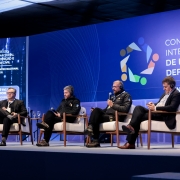 Quatro pessoas est&atilde;o em um palco, sentadas em cadeiras, uma delas fala ao microfone. Atr&aacute;s, um grande painel indicando Congresso Internacional de Prote&ccedil;&atilde;o e Defesa Civil