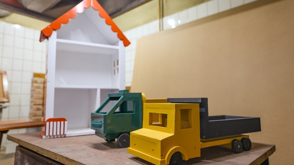 Mesa de madeira em ambiente interno com brinquedos artesanais, incluindo dois caminh&otilde;es coloridos e uma pequena estante em forma de casa com telhado vermelho.