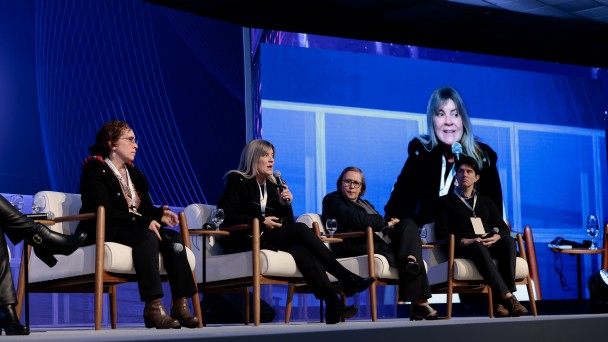 Esta &eacute; uma descri&ccedil;&atilde;o da imagem para fins de acessibilidade:

A fotografia em plano m&eacute;dio e &acirc;ngulo frontal-lateral registra a realiza&ccedil;&atilde;o de um painel de debates sobre um palco de eventos, identificado pelo t&iacute;tulo do arquivo como **"Painel Boas Praticas - foto Jo&atilde;o Pedro Rodrigues.jpg"**.

### **Os Integrantes do Painel**

* **Composi&ccedil;&atilde;o:** Quatro painelistas est&atilde;o sentados em uma fileira de poltronas brancas ou creme individuais com bra&ccedil;os e pernas de madeira clara, dispostas ao longo do palco. Todos usam credenciais oficiais suspensas por cord&otilde;es no pesco&ccedil;o.
* **Da esquerda para a direita:**
* Uma mulher de &oacute;culos e cabelos cacheados escuros, vestindo casaco preto e botas marrons de salto, est&aacute; sentada voltada de perfil parcial para a direita, segurando um microfone na m&atilde;o esquerda.
* Uma mulher de cabelos longos loiros ou claros est&aacute; centralizada no palco, falando ao microfone que segura com a m&atilde;o direita. Ela veste um casaco preto, cal&ccedil;a escura e botas pretas de salto.
* Uma mulher de &oacute;culos e cabelos curtos escuros est&aacute; sentada em pose reflexiva, acompanhando a fala da colega. Ela veste um casaco escuro de inverno e sapatos pretos.
* Um homem jovem de cabelos escuros e curtos est&aacute; sentado na extremidade direita do grupo, vestindo um su&eacute;ter preto sobre camisa social e cal&ccedil;a escura, com as m&atilde;os unidas sobre as pernas.


* **Extrema Esquerda:** Na borda esquerda inferior da imagem, veem-se apenas as pernas de outra participante vestindo cal&ccedil;a escura e botas de couro pretas de cano alto com fivelas douradas.

### **O Tel&atilde;o e o Cen&aacute;rio de Fundo**

* **O Grande Painel de LED:** Atr&aacute;s dos participantes, estende-se um imenso tel&atilde;o digital com ilumina&ccedil;&atilde;o e fundo predominantemente na cor azul-viva.
* **Conte&uacute;do Digital:** O lado esquerdo do tel&atilde;o exibe grafismos abstratos compostos por linhas finas e curvas que se cruzam de forma ritmada. O lado direito projeta em tamanho ampliado a transmiss&atilde;o ao vivo em v&iacute;deo (transmiss&atilde;o simult&acirc;nea) da painelista loira central enquanto ela realiza o seu pronunciamento.
* **Mobili&aacute;rio de Apoio:** Pequenas mesas redondas de madeira escura est&atilde;o posicionadas nas laterais e entre as poltronas, servindo de apoio para copos e ta&ccedil;as de &aacute;gua de vidro transparente. O teto do audit&oacute;rio, com placas quadradas escuras e sistemas de ilumina&ccedil;&atilde;o embutidos, aparece de forma discreta no topo da composi&ccedil;&atilde;o.
