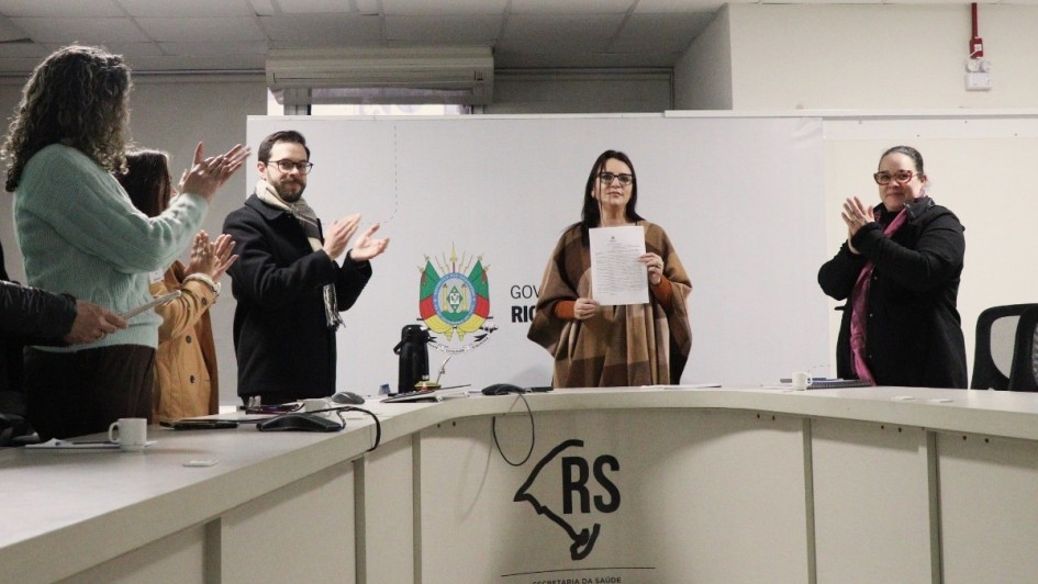 A fotografia em plano geral e &acirc;ngulo frontal registra o momento posterior &agrave; assinatura de um ato oficial em uma sala de reuni&otilde;es governamental. O t&iacute;tulo do arquivo identifica o contexto como a **"Assinatura da portaria que instituiu a Pol&iacute;tica Estadual de Monitoramento e Avalia&ccedil;&atilde;o em Sa&uacute;de 2026.jpeg"**.

### **As Autoridades e a A&ccedil;&atilde;o Central**

* **A Autoridade Central:** No centro do plano, atr&aacute;s de uma grande bancada, uma mulher de cabelos escuros e &oacute;culos de grau est&aacute; em p&eacute;, voltada para a frente. Ela segura com a m&atilde;o esquerda um documento oficial impresso em folha branca, exibindo-o para a c&acirc;mera. Ela veste uma blusa terracota sob um poncho de l&atilde; em tons de marrom e bege com estampa quadriculada.
* **Grupo de Apoio (&agrave; esquerda):**
* Um homem de &oacute;culos, barba e vestindo um casaco preto espesso com um cachecol de padr&atilde;o xadrez claro est&aacute; em p&eacute;, sorrindo e batendo palmas em dire&ccedil;&atilde;o &agrave; autoridade central.
* Ao lado dele, mais &agrave; esquerda, duas mulheres tamb&eacute;m aparecem em p&eacute; batendo palmas; uma veste um casaco caramelo e a outra, mais pr&oacute;xima &agrave; borda esquerda, veste um su&eacute;ter de tric&ocirc; verde-claro. Uma m&atilde;o masculina com casaco escuro aparece no canto inferior esquerdo segurando uma caneta.


* **Autoridade &agrave; Direita:** No lado direito da imagem, outra mulher de &oacute;culos com arma&ccedil;&atilde;o avermelhada e cabelos presos acompanha o ato em p&eacute;. Ela sorri e bate palmas, vestindo um casaco preto de inverno sobre uma echarpe ou blusa rosa vibrante.

### **O Ambiente e Elementos Institucionais**

* **A Bancada:** As autoridades est&atilde;o posicionadas atr&aacute;s de uma grande mesa ou bancada curva de trabalho em tom cinza-claro ou branco. Na face frontal dessa bancada, voltada para o espectador, destaca-se a aplica&ccedil;&atilde;o em preto do logotipo oficial estilizado do mapa do Rio Grande do Sul acompanhado da inscri&ccedil;&atilde;o **"RS / SECRETARIA DA SA&Uacute;DE"**. Sobre a mesa, veem-se x&iacute;caras de caf&eacute; brancas, pap&eacute;is, canetas, um mouse e uma garrafa t&eacute;rmica preta com uma cuia de chimarr&atilde;o ao centro.
* **O Painel de Fundo:** Atr&aacute;s das autoridades, h&aacute; um painel ou divis&oacute;ria branca port&aacute;til onde est&aacute; impresso o bras&atilde;o oficial do Estado do Rio Grande do Sul colorido, seguido pelas letras mai&uacute;sculas pretas **"GOV / RIO"** (com o restante do texto encoberto).
* **Cen&aacute;rio:** A sala possui teto de placas modulares brancas com lumin&aacute;rias embutidas e um aparelho de ar-condicionado instalado ao fundo. A ilumina&ccedil;&atilde;o interna &eacute; difusa e uniforme, concentrando a aten&ccedil;&atilde;o no grupo reunido para a celebra&ccedil;&atilde;o do ato.