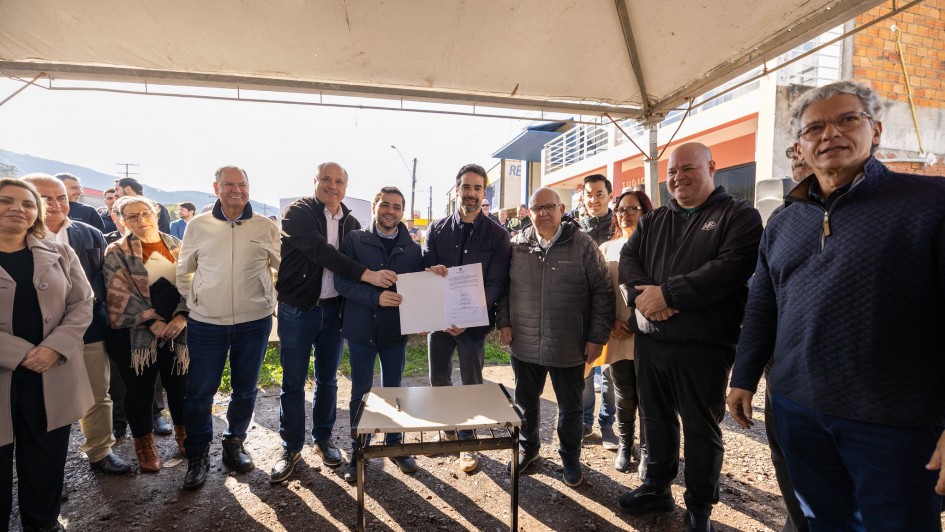 Esta &eacute; uma descri&ccedil;&atilde;o da imagem para fins de acessibilidade:

A fotografia em plano geral e &acirc;ngulo frontal registra o momento posterior &agrave; assinatura de um ato oficial ao ar livre. O t&iacute;tulo do arquivo contextualiza o evento: **"Governo do Estado assina conv&ecirc;nio para duplica&ccedil;&atilde;o da ERS-030 em Os&oacute;rio.jpg"**.

### **As Autoridades e a A&ccedil;&atilde;o Central**

* **O Grupo Central:** No centro da imagem, sob a cobertura de uma tenda, um grupo de autoridades pol&iacute;ticas e representantes p&uacute;blicos est&aacute; perfilado lado a lado, sorrindo em dire&ccedil;&atilde;o &agrave; c&acirc;mera.
* **Exibi&ccedil;&atilde;o do Documento:** O governador do estado est&aacute; posicionado ao centro-direita do grupo, vestindo uma jaqueta escura sobre camiseta escura. Ele segura, junto com o homem de jaqueta azul-marinho &agrave; sua esquerda, uma pasta branca aberta contendo o documento oficial assinado, exibindo-o frontalmente.
* **Demais Integrantes:**
* &Agrave; esquerda do homem de jaqueta azul, um homem de jaqueta preta aberta sobre camisa social clara estende o bra&ccedil;o em dire&ccedil;&atilde;o ao documento. Ao lado deste, um homem de jaqueta bege clara e cal&ccedil;a jeans acompanha o ato sorridente. Na extrema esquerda, veem-se uma mulher com casaco sobretudo claro e outras pessoas ao fundo.
* &Agrave; direita do governador, um homem idoso de &oacute;culos usa uma jaqueta acolchoada cinza escuro. Ao seu lado, destacam-se um homem calvo de jaqueta preta esportiva com os bra&ccedil;os cruzados &agrave; frente e, na extrema direita em primeiro plano, um homem de &oacute;culos e cabelos grisalhos vestindo um su&eacute;ter texturizado azul-marinho.



### **O Mobili&aacute;rio e o Ambiente**

* **A Mesa de Assinatura:** Centralizada em primeiro plano, logo &agrave; frente das autoridades, h&aacute; uma pequena mesa retangular simples com tampo claro e estrutura de metal preto, onde repousa uma caneta escura utilizada para as assinaturas.
* **O Cen&aacute;rio ao Redor:** O grupo est&aacute; posicionado sobre um terreno de terra batida e brita. Eles est&atilde;o abrigados sob uma grande tenda de lona clara, cuja estrutura met&aacute;lica e cobertura aparecem no topo da imagem.
* **O Plano de Fundo:** Ao fundo, sob a luz forte do sol, avistam-se edif&iacute;cios urbanos, incluindo um pr&eacute;dio de dois pavimentos com sacada de grade branca e uma parede de tijolos sem reboco &agrave; direita. Elementos como postes de energia com fia&ccedil;&atilde;o a&eacute;rea e a silhueta de morros distantes na extrema esquerda completam a paisagem externa.