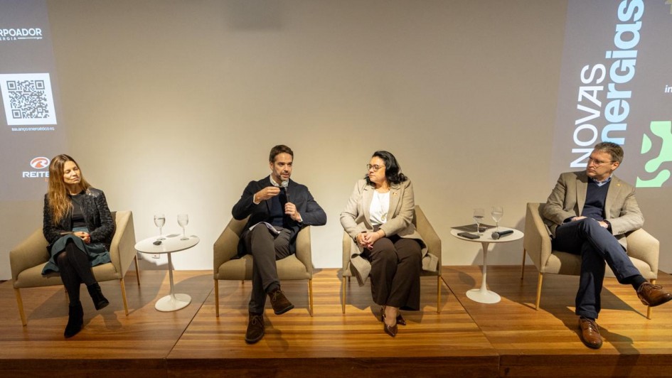 Vis&atilde;o frontal do painel com quatro participantes sentados lado a lado em poltronas sobre o palco, com pequenas mesas de apoio entre eles e proje&ccedil;&otilde;es laterais com elementos gr&aacute;ficos e as palavras &ldquo;Novas Energias&rdquo;.