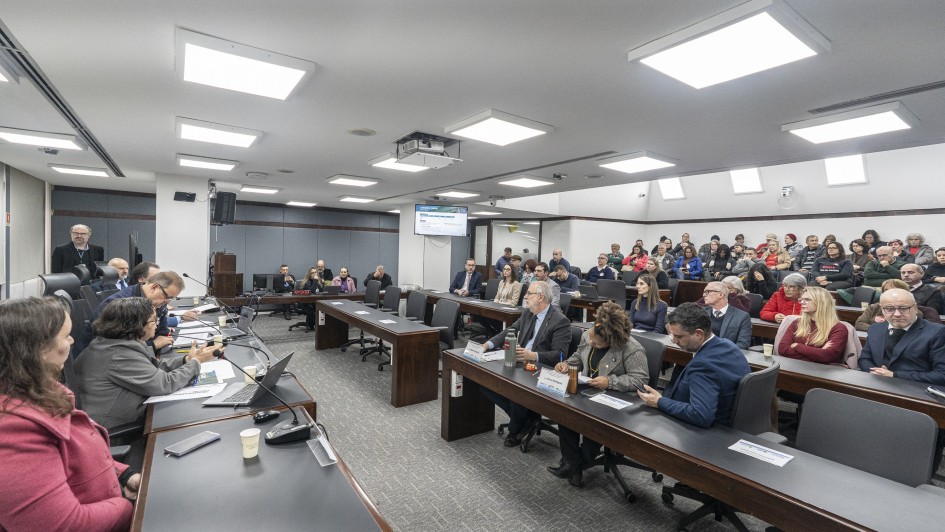 Sala ampla de reuni&atilde;o institucional com mesas longas, onde diversas pessoas est&atilde;o sentadas acompanhando uma apresenta&ccedil;&atilde;o; &agrave; frente, um grupo utiliza microfones e computadores, enquanto no centro da sala h&aacute; um projetor e um monitor exibindo conte&uacute;do t&eacute;cnico. 