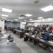 Sala ampla de reuni&atilde;o institucional com mesas longas, onde diversas pessoas est&atilde;o sentadas acompanhando uma apresenta&ccedil;&atilde;o; &agrave; frente, um grupo utiliza microfones e computadores, enquanto no centro da sala h&aacute; um projetor e um monitor exibindo conte&uacute;do t&eacute;cnico. 