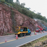 Trecho de rodovia em obra com cones sinalizando a pista, onde caminh&otilde;es e equipamentos operam junto a um pared&atilde;o rochoso; ao lado, um ve&iacute;culo de apoio com estrutura met&aacute;lica transporta materiais, enquanto trabalhadores atuam pr&oacute;ximos ao talude em meio &agrave; vegeta&ccedil;&atilde;o.