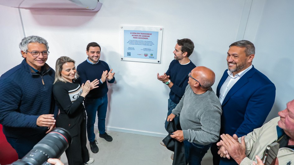 Espa&ccedil;o fechado onde um grupo de pessoas est&aacute; reunido ao redor de uma placa comemorativa fixada na parede, com o texto &ldquo;A vida em Tramanda&iacute; acaba de ganhar mais agilidade&rdquo;.