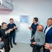 Espa&ccedil;o fechado onde um grupo de pessoas est&aacute; reunido ao redor de uma placa comemorativa fixada na parede, com o texto &ldquo;A vida em Tramanda&iacute; acaba de ganhar mais agilidade&rdquo;.