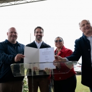 Momento de registro ap&oacute;s a assinatura, com quatro pessoas exibindo um documento aberto sobre o p&uacute;lpito para a c&acirc;mera, em um ambiente externo com gramado e participantes ao redor.