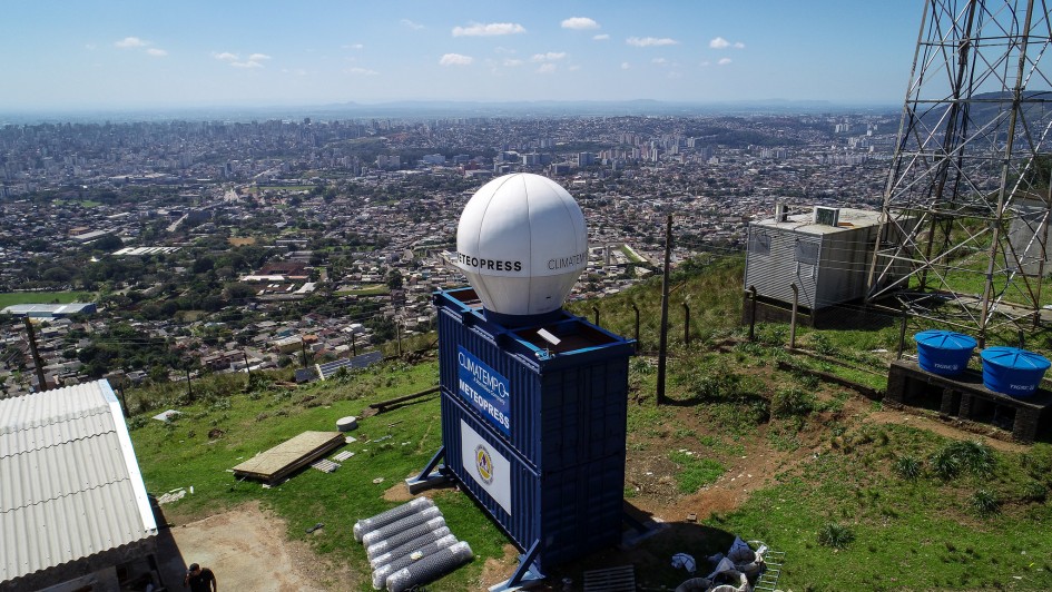 Vista elevada de uma &aacute;rea urbana extensa, tendo em primeiro plano uma estrutura de monitoramento meteorol&oacute;gico instalada sobre um cont&ecirc;iner azul; ao lado h&aacute; uma torre met&aacute;lica de telecomunica&ccedil;&otilde;es, e ao fundo se espalha a paisagem da cidade sob c&eacute;u azul com poucas nuvens.