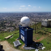 Vista elevada de uma &aacute;rea urbana extensa, tendo em primeiro plano uma estrutura de monitoramento meteorol&oacute;gico instalada sobre um cont&ecirc;iner azul; ao lado h&aacute; uma torre met&aacute;lica de telecomunica&ccedil;&otilde;es, e ao fundo se espalha a paisagem da cidade sob c&eacute;u azul com poucas nuvens.