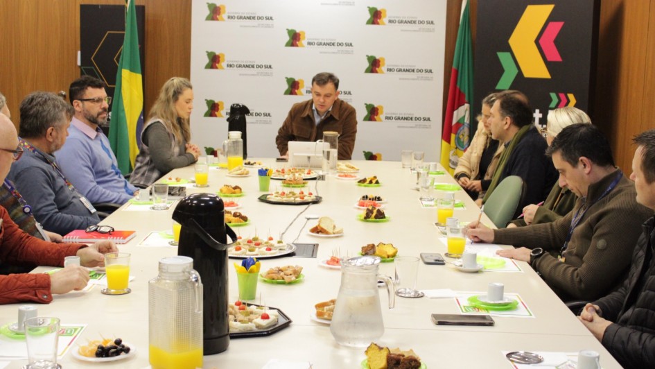 Aqui est&aacute; a descri&ccedil;&atilde;o da imagem para fins de acessibilidade 
---

**Descri&ccedil;&atilde;o da imagem "Governo do Estado se re&uacute;ne com entidades do varejo para preparar Ganha-Ganha Farroupilha 2026.jpg":**

Uma fotografia colorida em plano m&eacute;dio mostra uma reuni&atilde;o de trabalho em torno de uma grande mesa retangular branca em uma sala de confer&ecirc;ncias. Cerca de dez pessoas participam do encontro, distribu&iacute;das ao longo das laterais e na cabeceira da mesa ao fundo.

**Disposi&ccedil;&atilde;o dos participantes:**

* **Lado esquerdo da mesa:** H&aacute; quatro pessoas sentadas de perfil parcial. Em primeiro plano, v&ecirc;-se apenas a m&atilde;o e o bra&ccedil;o de um homem com casaco avermelhado. Ao lado dele, um homem de blusa cinza-escura e crach&aacute; institucional. Em seguida, um homem de &oacute;culos e barba vestindo uma camisa social azul-clara e, mais ao fundo, uma mulher de cabelos loiros longos vestindo um colete quadriculado cinza.
* **Cabeceira da mesa (ao fundo):** Um homem de cabelos escuros curtos est&aacute; sentado no centro, vestindo uma jaqueta de camur&ccedil;a ou veludo marrom sobre uma camisa azul-clara. Ele est&aacute; levemente inclinado para a frente, olhando para um notebook aberto &agrave; sua frente.
* **Lado direito da mesa:** H&aacute; cinco pessoas sentadas. Mais ao fundo, uma mulher de casaco bege, seguida por um homem com cachecol verde-oliva. Ao centro, uma mulher loira de &oacute;culos e, em primeiro plano, dois homens com casacos escuros focados na reuni&atilde;o, estando um deles escrevendo em pap&eacute;is com uma caneta.

**Elementos sobre a mesa:**
A mesa est&aacute; farta com itens de caf&eacute; da manh&atilde; ou *coffee break*. H&aacute; jarras de vidro com suco de laranja, garrafas t&eacute;rmicas pretas para caf&eacute; ou &aacute;gua, copos de vidro e x&iacute;caras de caf&eacute; sobre pires com detalhes verdes. Tamb&eacute;m est&atilde;o distribu&iacute;dos diversos pratinhos e bandejas com alimentos, incluindo peda&ccedil;os de bolo, frutas picadas (como melancia e uvas), sandu&iacute;ches em miniatura com palitos e salgados fritos.

**Ambiente e plano de fundo:**
As paredes laterais possuem revestimento de pain&eacute;is de madeira marrom-clara. Ao fundo da sala, atr&aacute;s do homem na cabeceira, destaca-se um grande painel vertical branco com repeti&ccedil;&otilde;es do logotipo oficial do **"GOVERNO DO ESTADO RIO GRANDE DO SUL - SECRETARIA DE DESENVOLVIMENTO ECON&Ocirc;MICO"**. Ladeando esse painel, h&aacute; duas bandeiras oficiais em pedestais (incluindo a do Brasil &agrave; esquerda e a do Rio Grande do Sul &agrave; direita) e um banner vertical preto no canto superior direito com a identidade visual da marca **"invest.RS"**, exibindo formas geom&eacute;tricas em amarelo, verde e rosa.