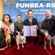 Grupo de quatro pessoas posando sobre um tapete vermelho em frente a um painel com o texto &ldquo;FUNBEA-RS&rdquo;, exibindo um documento aberto para a c&acirc;mera; tr&ecirc;s c&atilde;es aparecem em primeiro plano ao lado dos participantes.