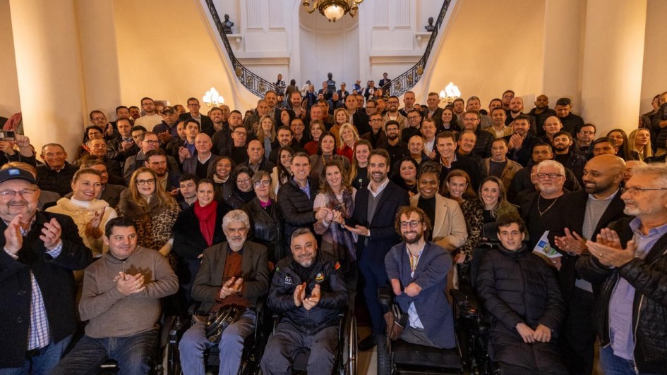 Grupo numeroso reunido no hall de um pr&eacute;dio hist&oacute;rico; v&aacute;rias pessoas ocupam o espa&ccedil;o central e os degraus, enquanto algumas utilizam cadeiras de rodas.